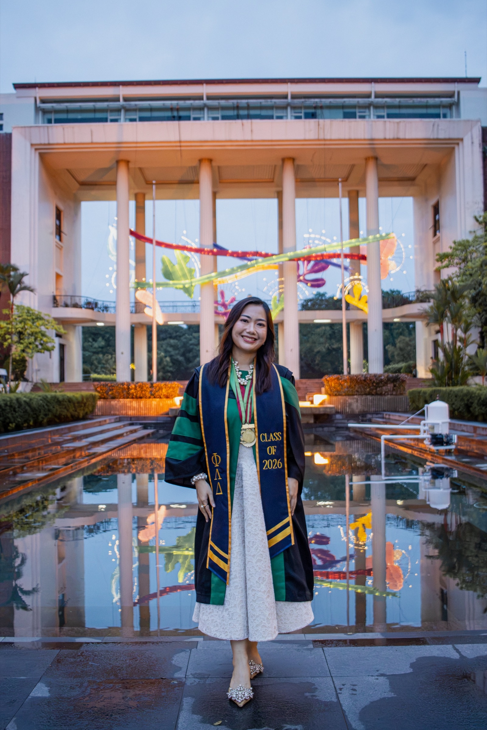 A graduate in full regalia stands before a lit hall at dusk, mirrored in the wet pavement