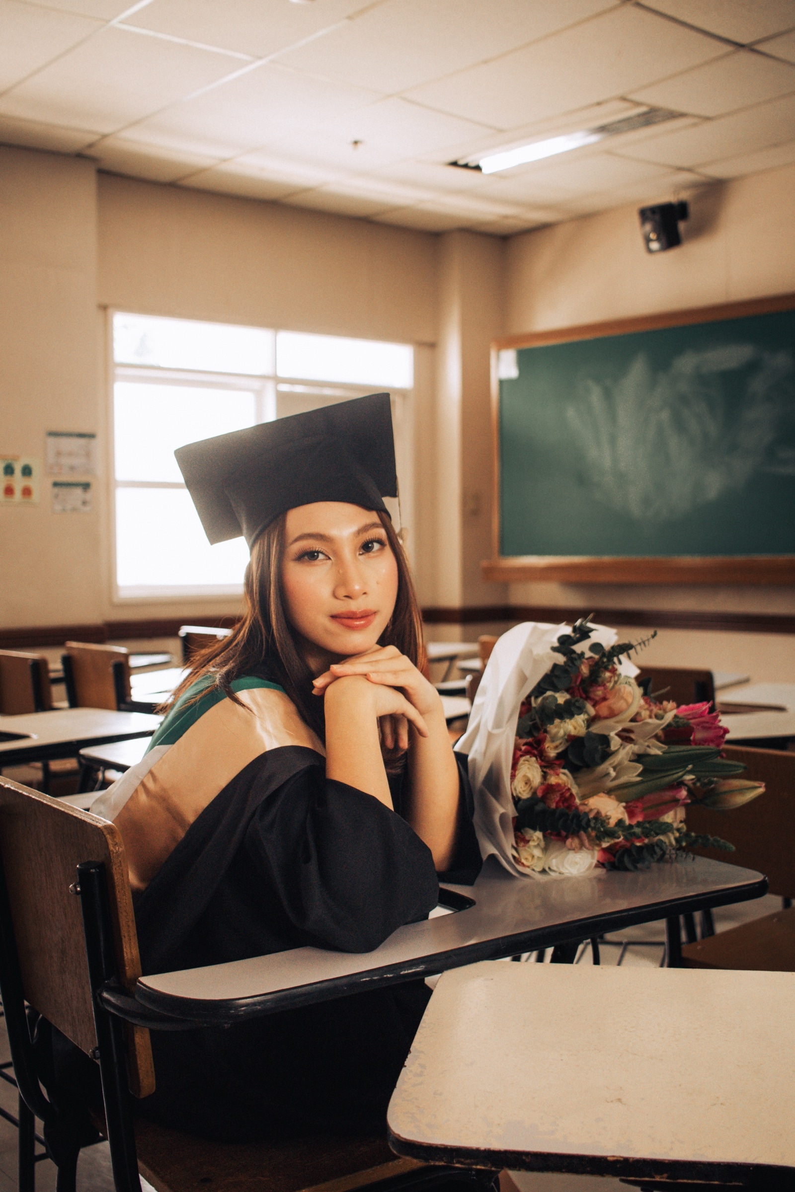 A graduate rests her chin on her hand at a classroom desk, a bouquet beside her