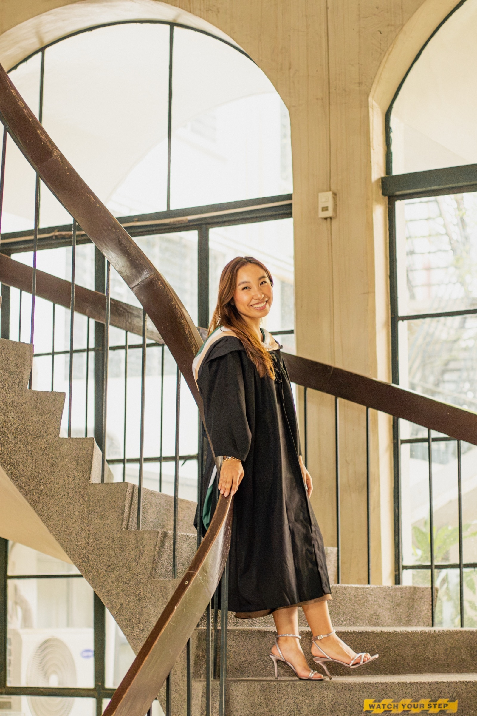 A graduate pauses on a spiral staircase beside tall arched windows