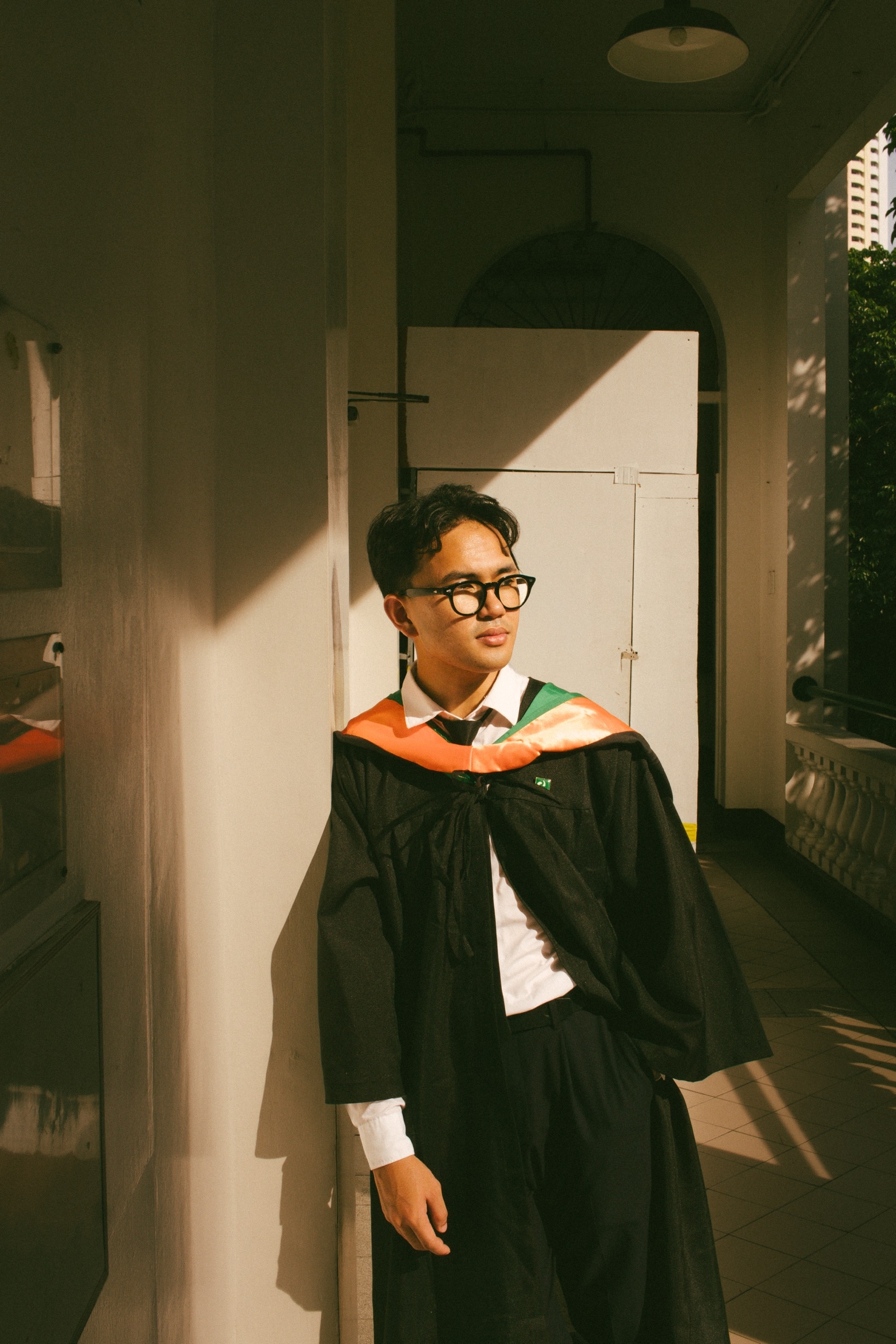 A graduate leans on a corridor wall in a strip of late afternoon sun
