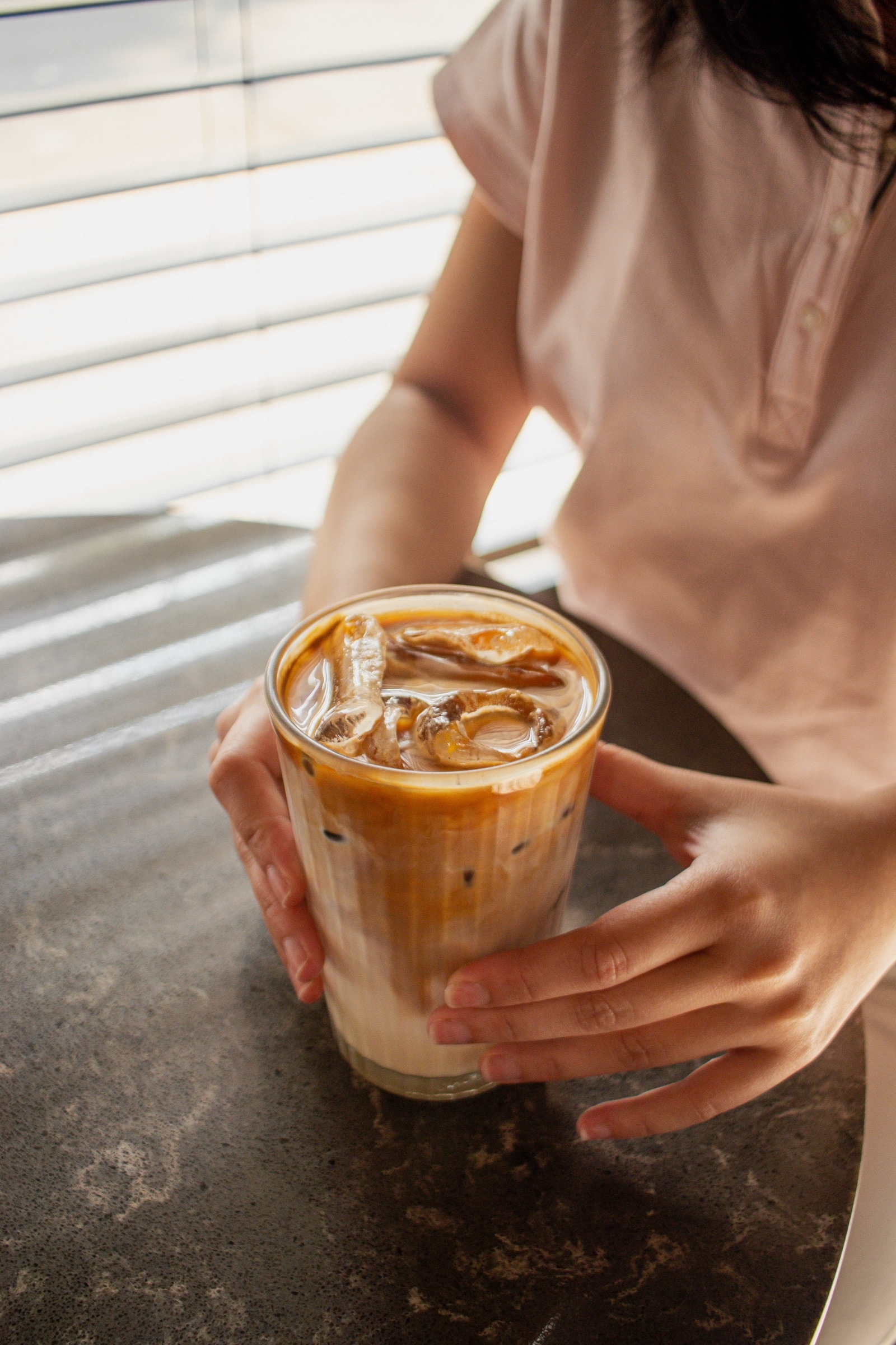 Two hands hold an iced latte on a stone table in striped window light