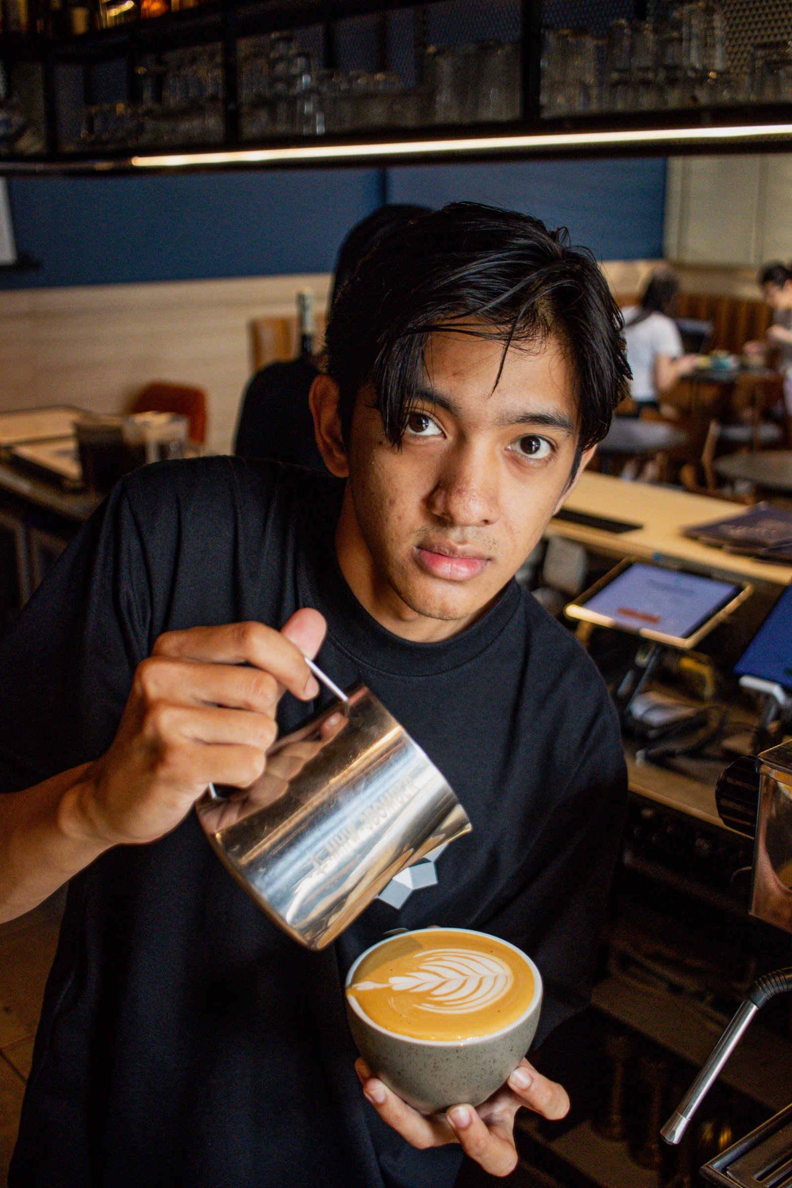 A barista pours milk into a latte and looks straight into the lens